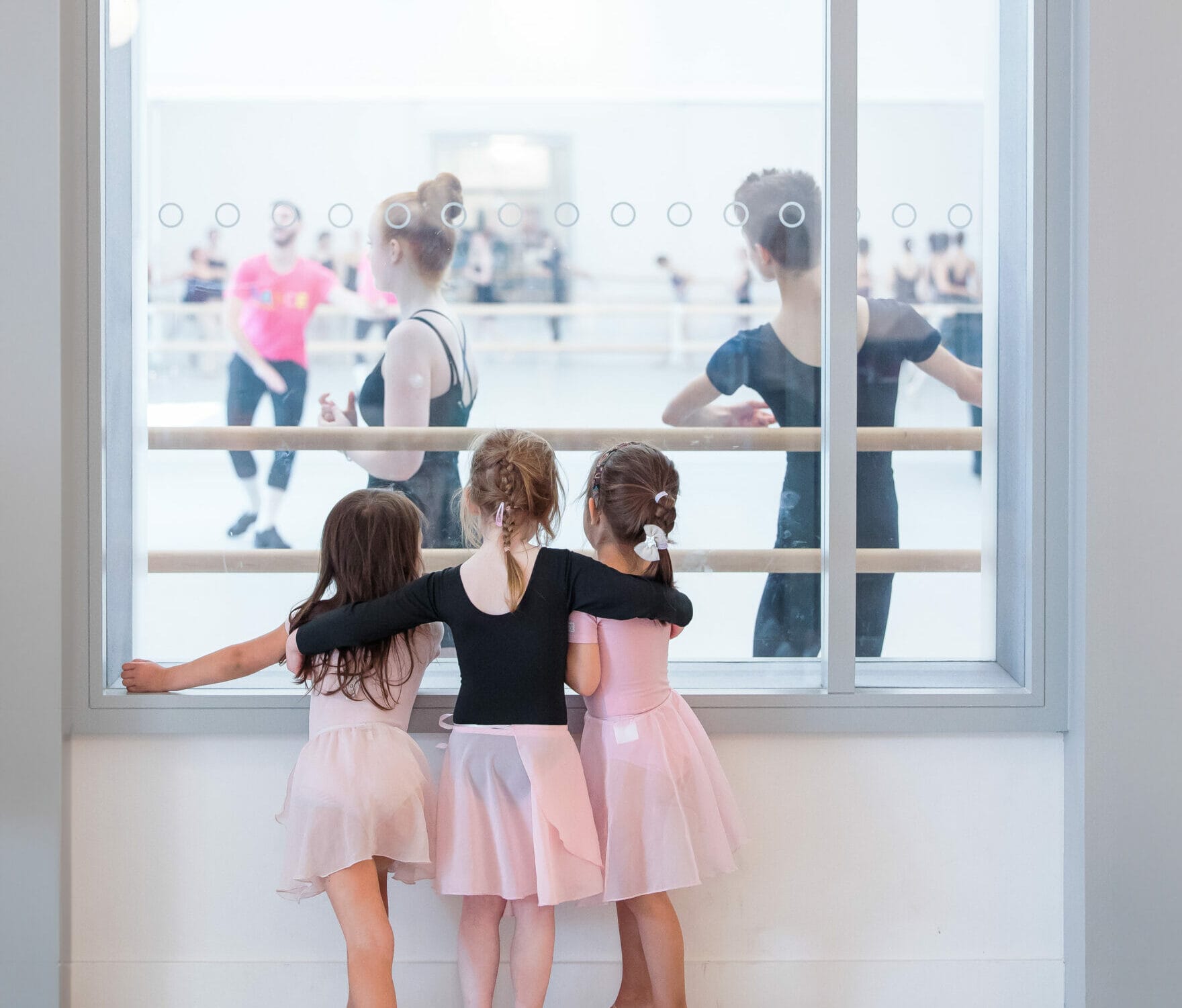 Image of three young children looking through window into older dance class for inspiration